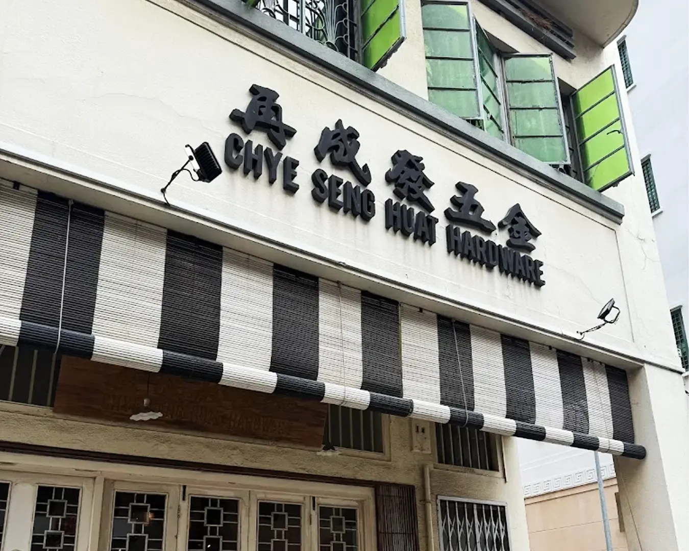 Facade of Chye Seng Huat Hardware store with black letters and Chinese characters. Black and white striped awning; open green window shutters above.