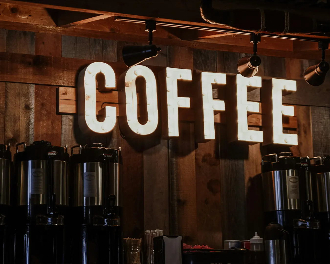 Illuminated "COFFEE" sign on a rustic wooden wall, with several coffee dispensers below, creating a warm and inviting café ambiance.