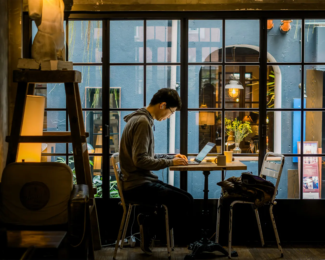 A person in a hoodie works on a laptop in a cozy café, next to a window with a warm, artsy ambiance. Soft lighting and indoor plants set a calm tone.