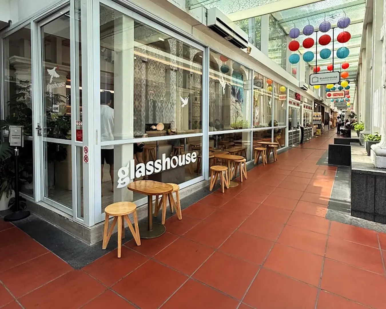 Corridor scene with glasshouse cafe on the left. Red tiled floor, wooden stools, and colorful hanging lanterns create a vibrant, inviting atmosphere.