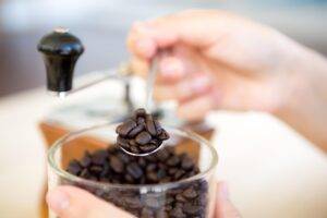 A person uses a small spoon to scoop dark roasted coffee beans from a clear glass jar. In the blurred background, a traditional manual coffee grinder sits ready for the brewing process.