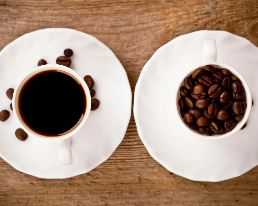 Two white cups on saucers on a wooden surface; one filled with black coffee, the other filled with roasted coffee beans.