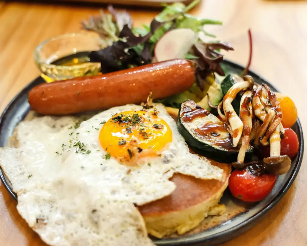 A hearty breakfast plate with a sunny-side-up egg, sausage, pancake, grilled veggies, assorted greens, and a small bowl of syrup on a wooden table.