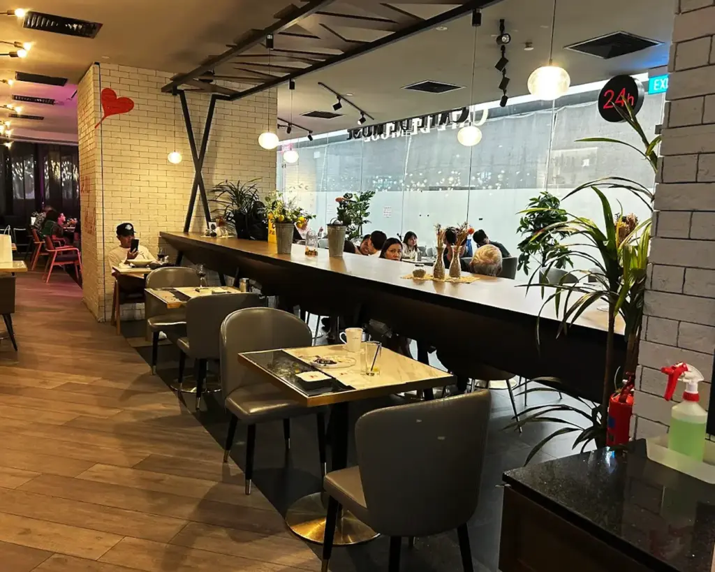 Cozy café interior with wooden floors, brick walls, and pendant lights. Tables and chairs are set, with plants and a 24-hour sign visible. A relaxed, warm atmosphere with people enjoying their meals.