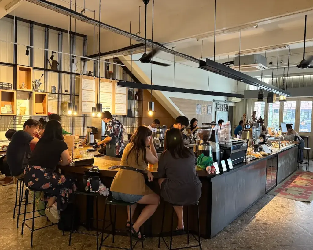 Spacious café with a warm ambiance, featuring people seated at the counter and baristas preparing drinks. Natural light streams through large windows.