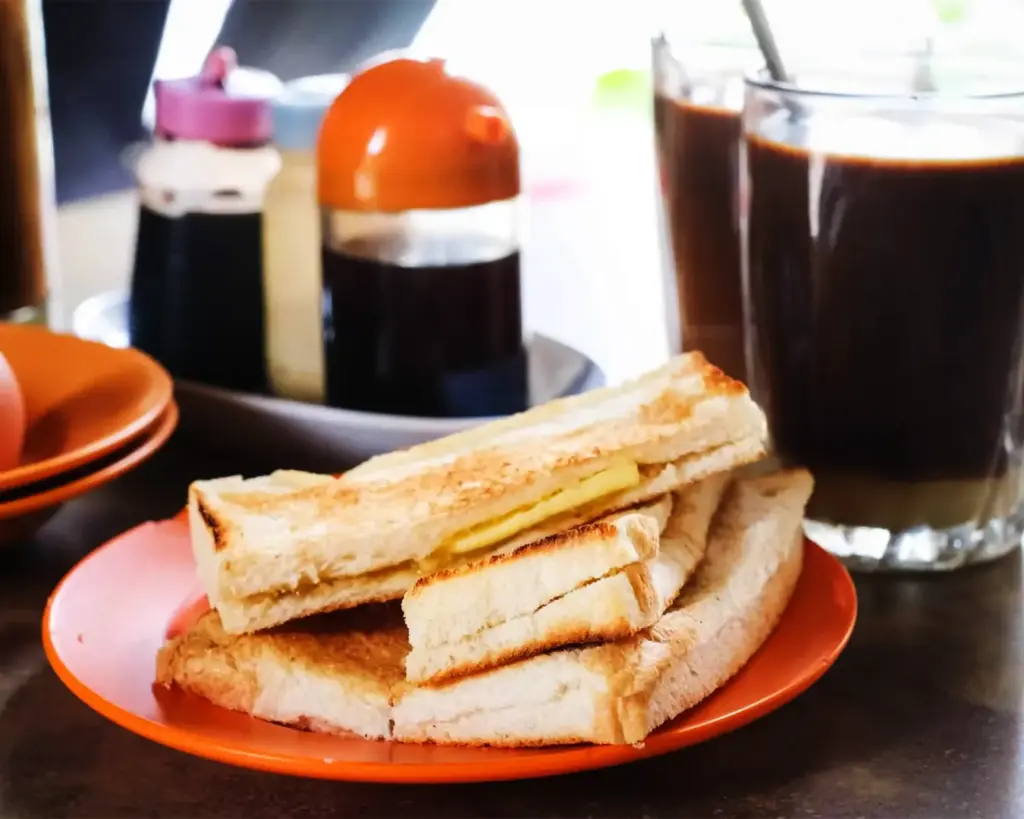 Toasted bread slices with butter on an orange plate, next to a glass of dark drink. Background has condiments, creating a cozy breakfast scene.