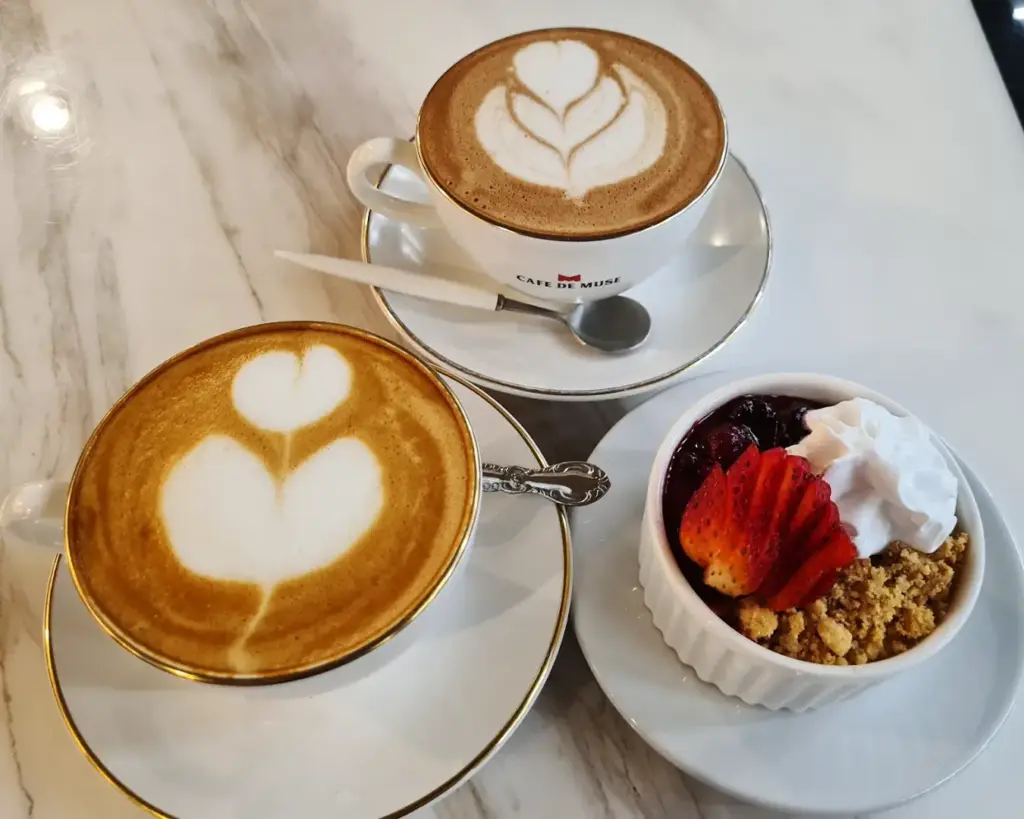 Two cappuccinos with heart-shaped latte art on a marble table beside a small bowl of dessert with strawberries, berries, whipped cream, and crumbles.