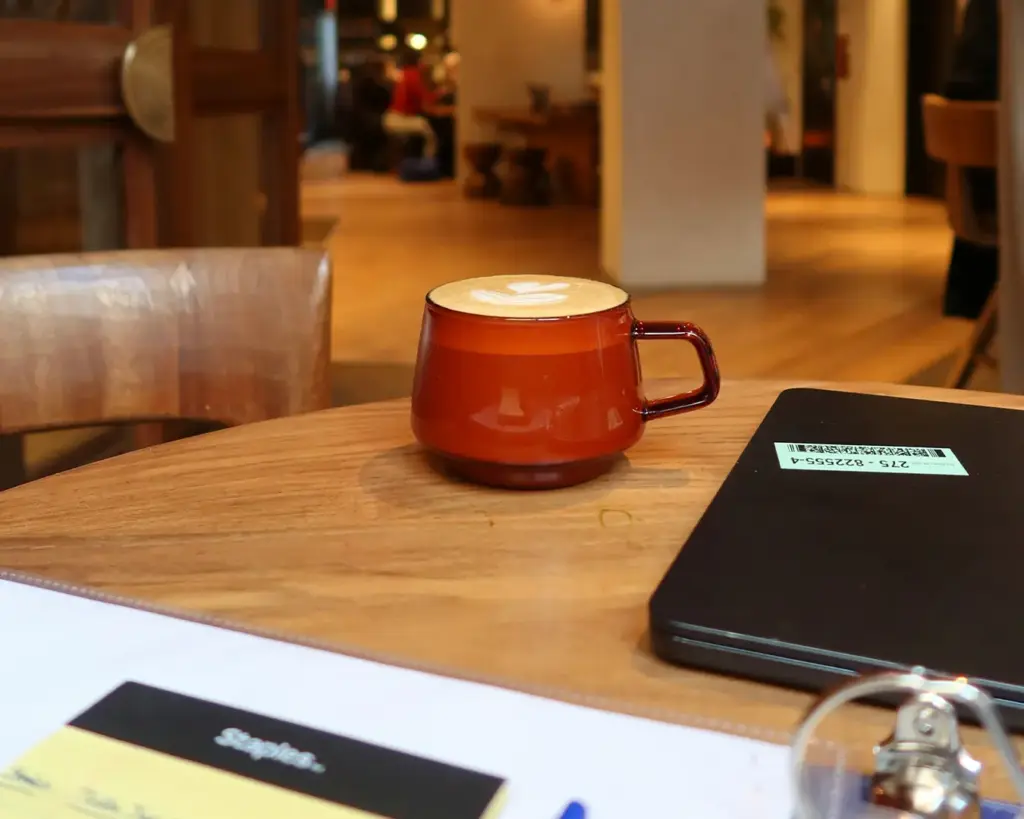 A cozy café scene with a warm wooden table featuring a red mug filled with a latte, next to a closed black notebook. The background is softly blurred, with people seated, creating a relaxed atmosphere.