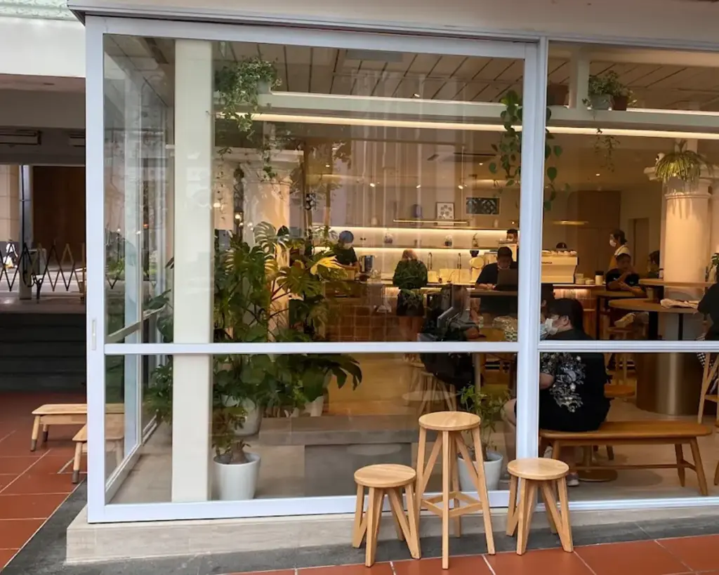 Cozy cafe interior seen through large window, with wooden stools outside. Inside, people sit at tables amid lush green plants and warm lighting.