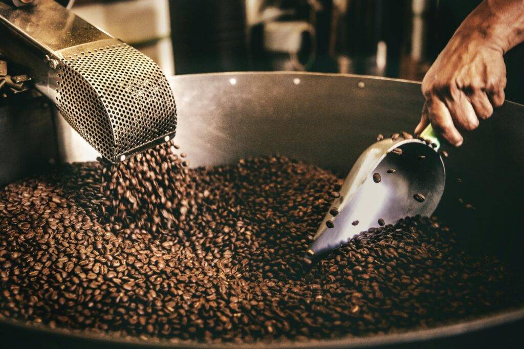 Freshly roasted coffee beans cascade into a large cooling tray while a metal scoop stirs through the warm, fragrant pile. The industrial setting and moody lighting highlight the rich textures of the beans during the roasting process.