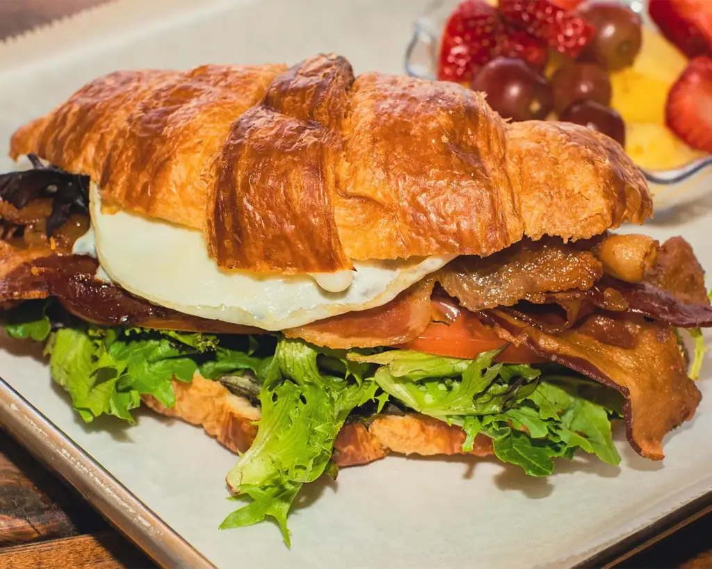 A flaky croissant sandwich filled with crisp bacon, fried egg, fresh lettuce, and tomato. Served on a tray with a side of strawberries and grapes.