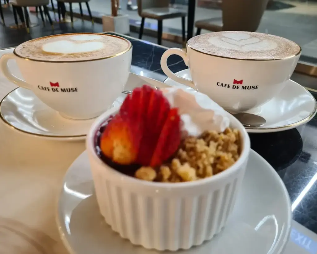 Two cappuccinos with latte art in branded cups sit on saucers at a café. A dessert with strawberries and whipped cream in a white ramekin is in the foreground.