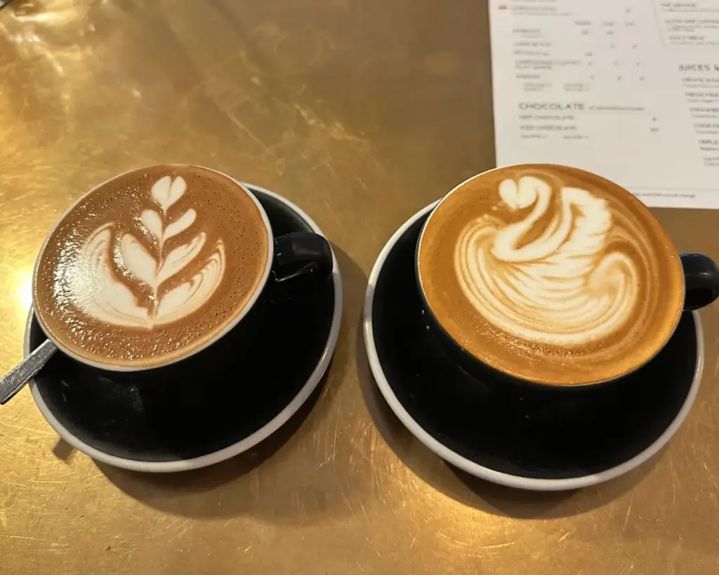 Two cappuccinos on a gold table; left cup features a leafy latte art; right cup displays a swan. Elegantly served in black saucers with a menu nearby.