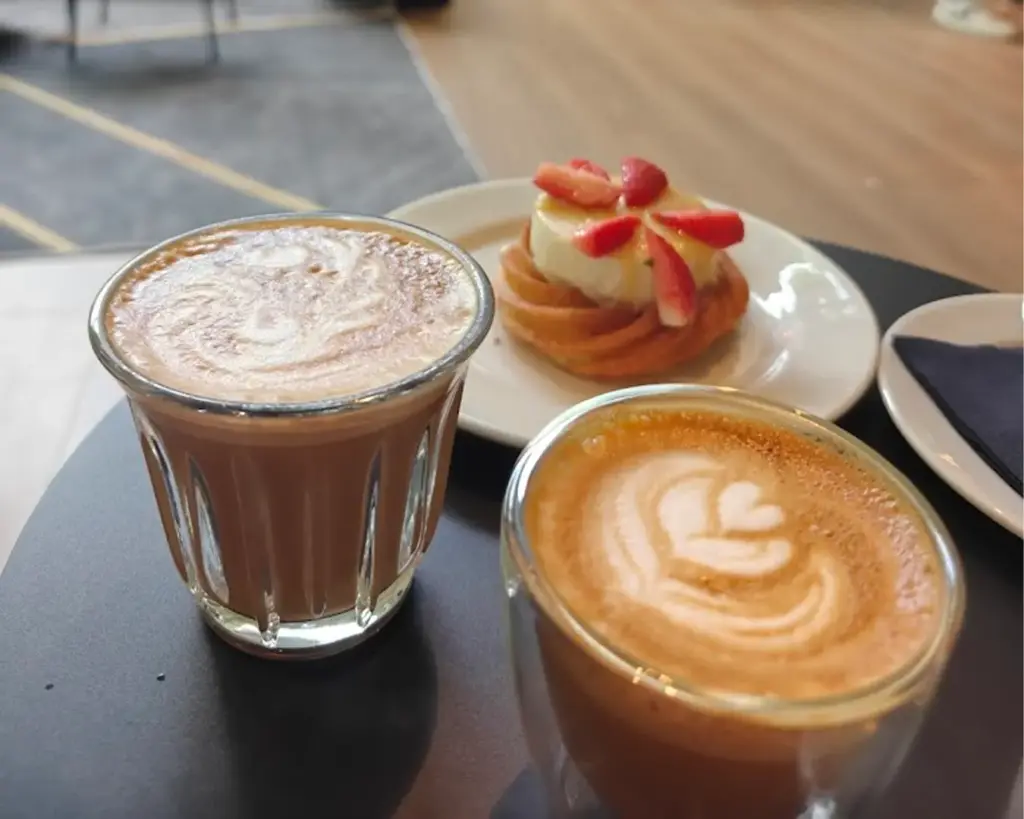 Two lattes with latte art sit on a table next to a pastry topped with cream and strawberries on a plate, creating a cozy cafe vibe.