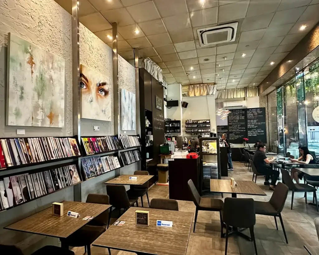 Cozy café interior with warm lighting. Walls display abstract art and shelves of magazines. Several tables, some occupied, create a relaxed, inviting atmosphere.