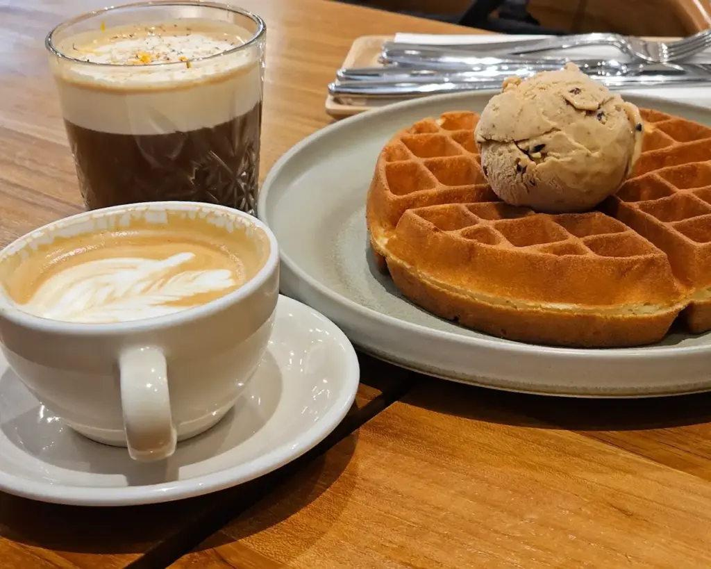 A latte in a ceramic cup and a glass of layered coffee sit next to a plate of waffles topped with a scoop of ice cream, on a wooden table.