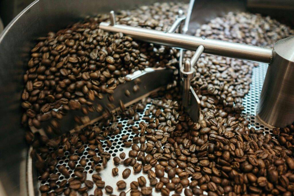 Freshly roasted coffee beans swirl within the cooling tray of a large industrial roasting machine. The metal arms of the roaster stir the dark brown beans over a perforated surface to ensure an even and efficient cooling process.