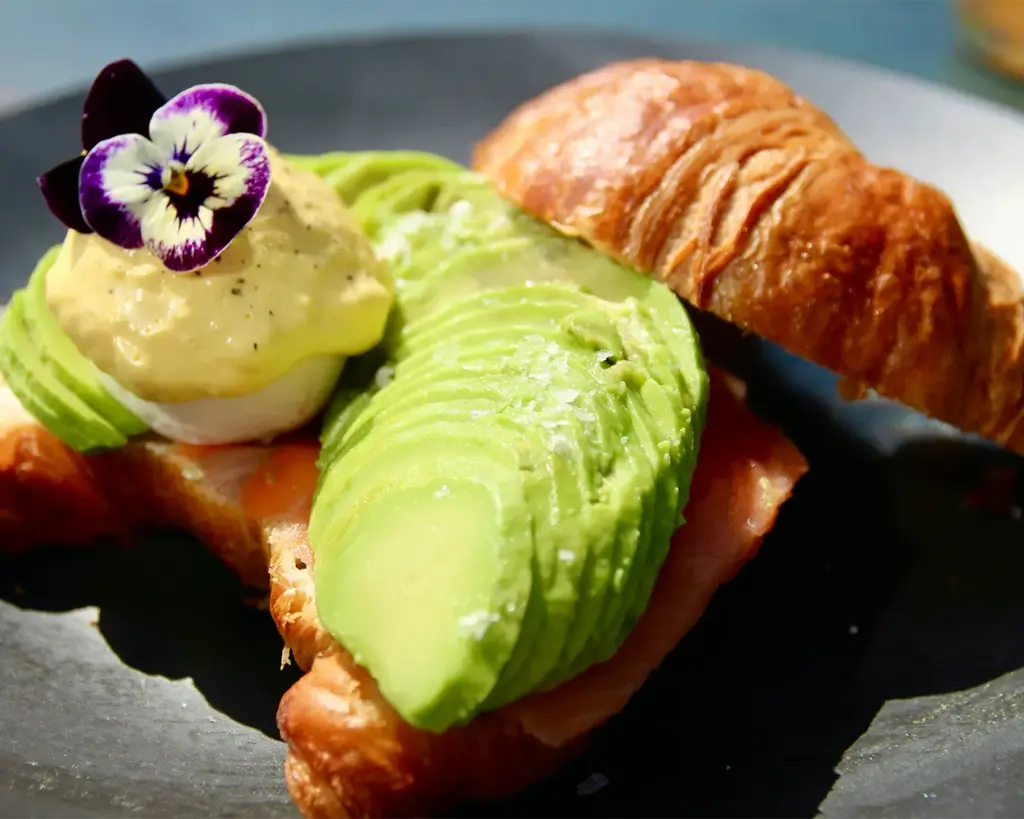 A croissant sandwich with sliced avocado, a poached egg topped with hollandaise sauce, and a purple pansy flower on a dark plate.