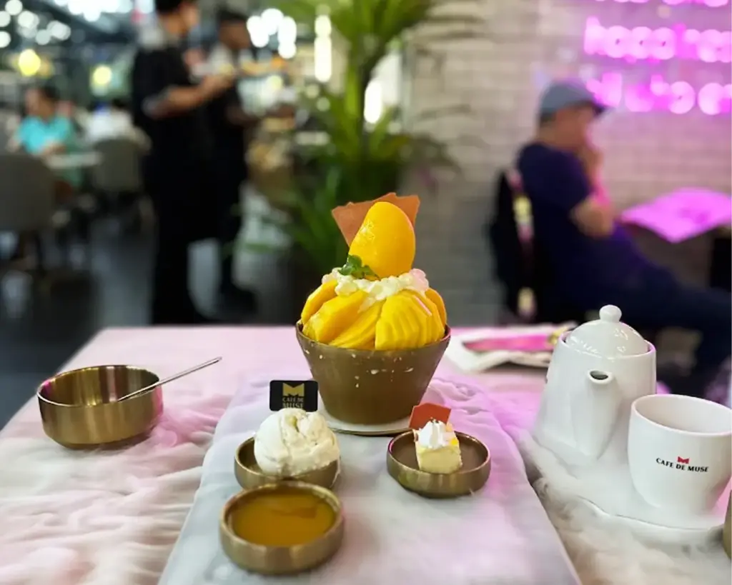 A vibrant dessert table with a mango-topped shaved ice dish, whipped cream, and syrup in bowls. A teapot and cup are set beside on a pink tablecloth.