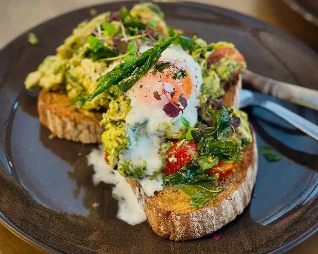 Avocado toast topped with a poached egg, greens, and cherry tomatoes on a dark plate. The dish looks fresh and flavorful, garnished with herbs.