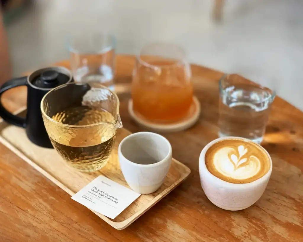 A cozy cafe setup on a wooden table featuring a latte with heart-shaped latte art, a glass of tea, an empty cup, an orange drink, and a glass of water.