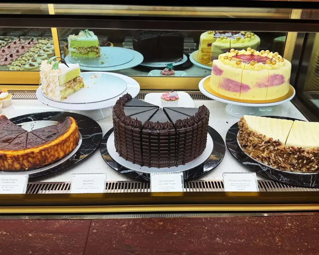A display case with assorted cakes: chocolate with layers, cheesecake, fruit-topped, and nut-coated. Colorful and appetizing presentation.