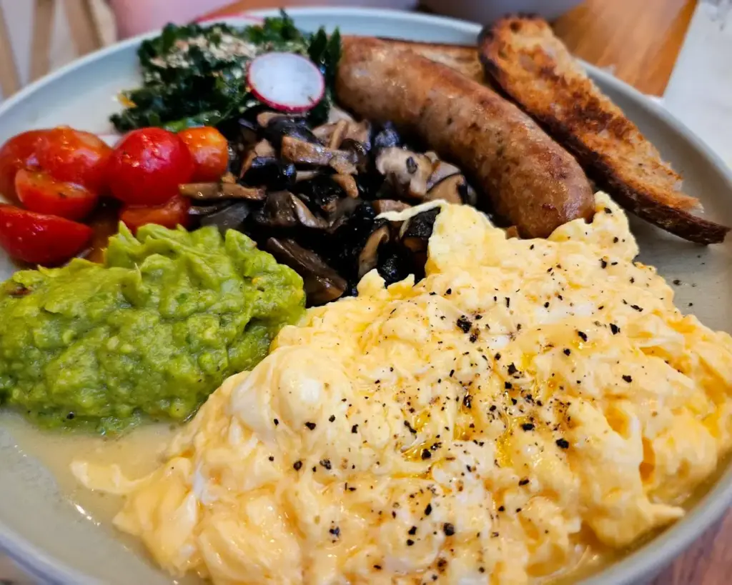 A hearty breakfast plate with creamy scrambled eggs, guacamole, sautéed mushrooms, roasted tomatoes, a sausage, kale, and toasted bread.