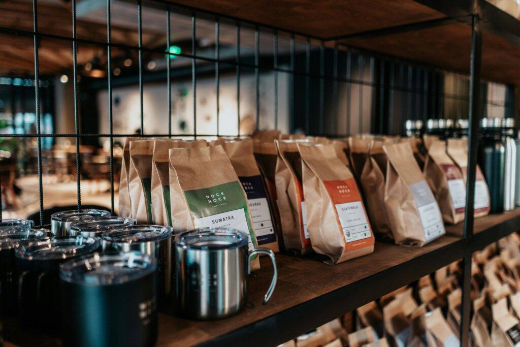 Rows of brown paper coffee bags with colorful labels, such as "Sumatra" and "Guatemala," are neatly arranged on a wooden shelf behind a black wire grid. In the foreground, sleek stainless steel and matte black travel mugs are lined up, reflecting the warm, industrial lighting of the modern cafe interior.