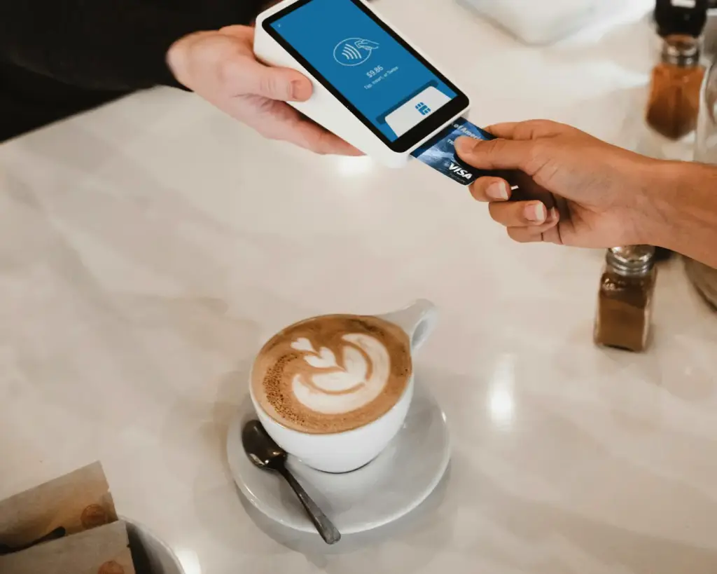A person paying with a credit card using a contactless card reader at a table with a cup of latte art coffee.