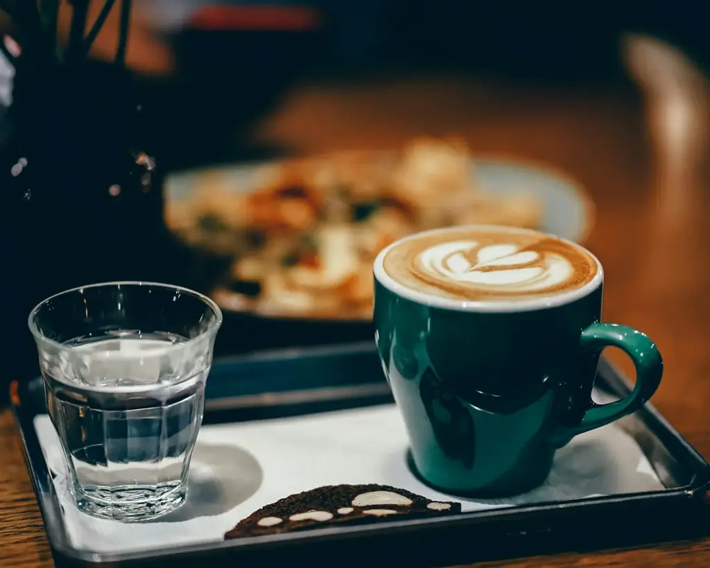 A green mug of latte with heart-shaped foam art sits on a tray alongside a glass of water and a slice of bread, set against a cozy cafe backdrop.