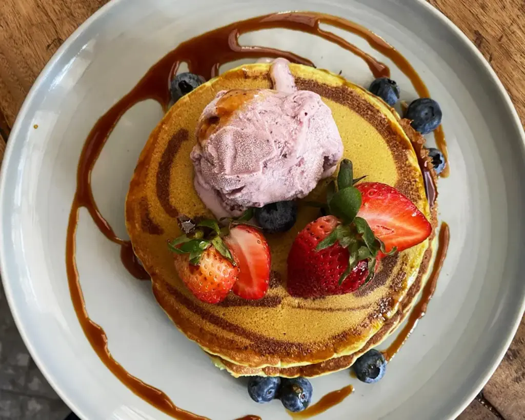 Pancakes topped with pink ice cream, fresh strawberries, and blueberries on a gray plate. Caramel sauce drizzled around the edges.