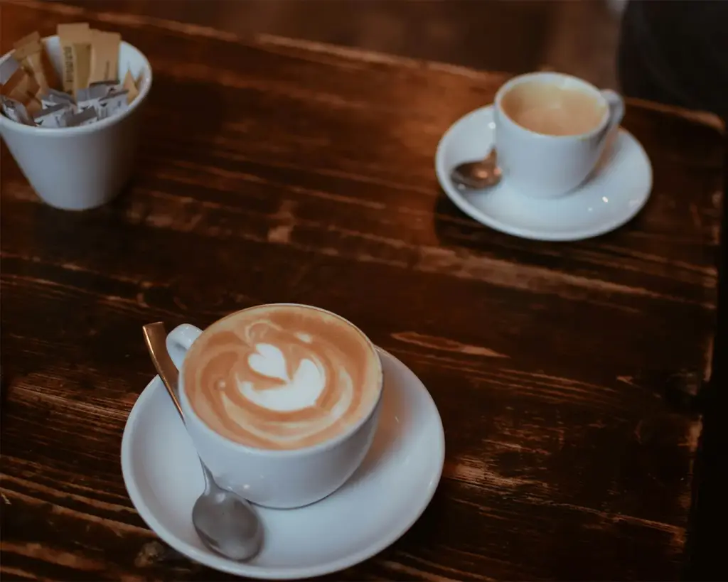 Two cups of coffee on a dark wooden table, one with heart latte art. A spoon rests on each saucer. Sugars in a cup nearby. Cozy and inviting ambiance.
