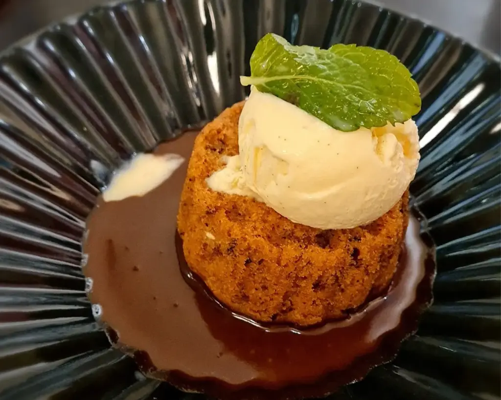 Warm cake topped with vanilla ice cream and a mint leaf, served on a glossy black plate with a pool of rich, dark chocolate sauce. Decadent and inviting.