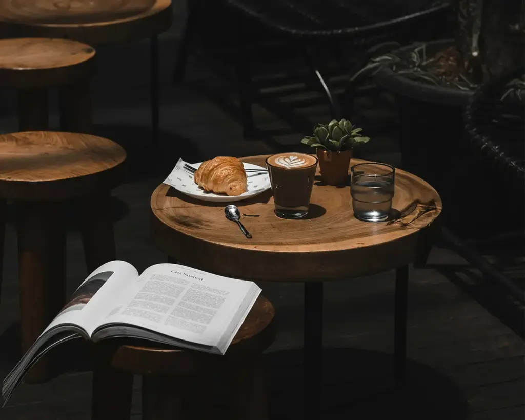 Cozy setup with a wooden table holding a croissant, a latte, a glass of water, and a small plant. An open book is on a nearby stool, creating a relaxed vibe.