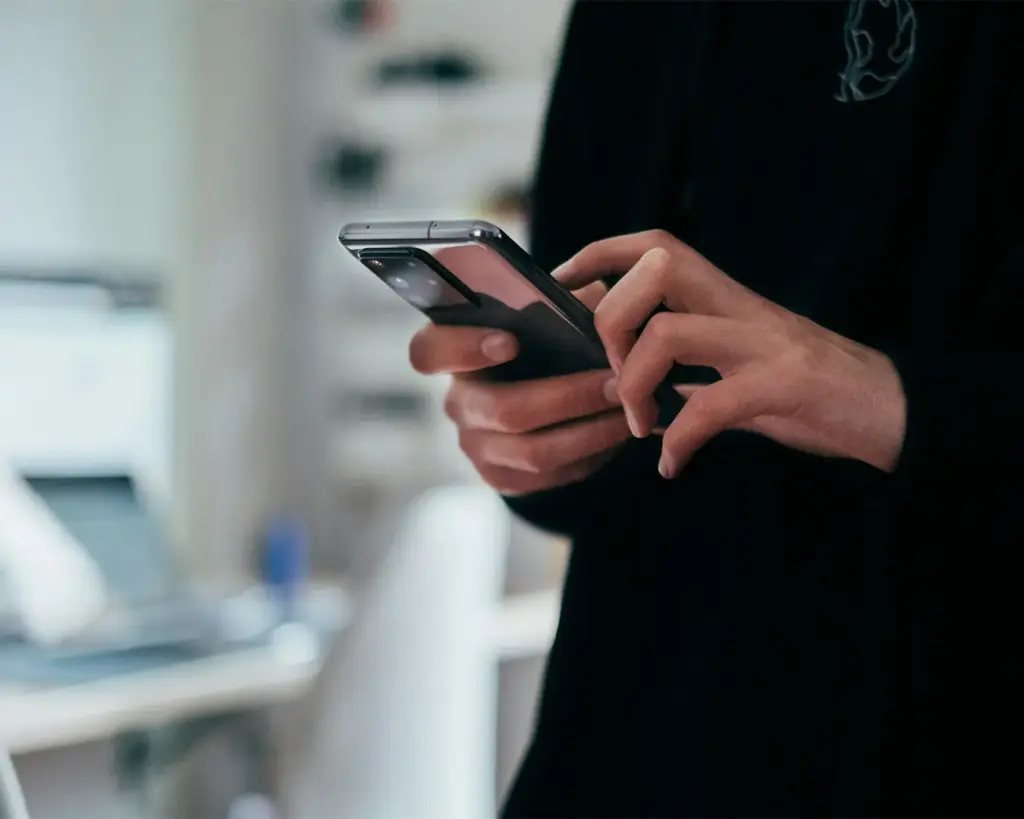A person in a dark shirt uses a smartphone indoors. The background is blurred, suggesting a modern, casual work or home setting. The mood is focused.