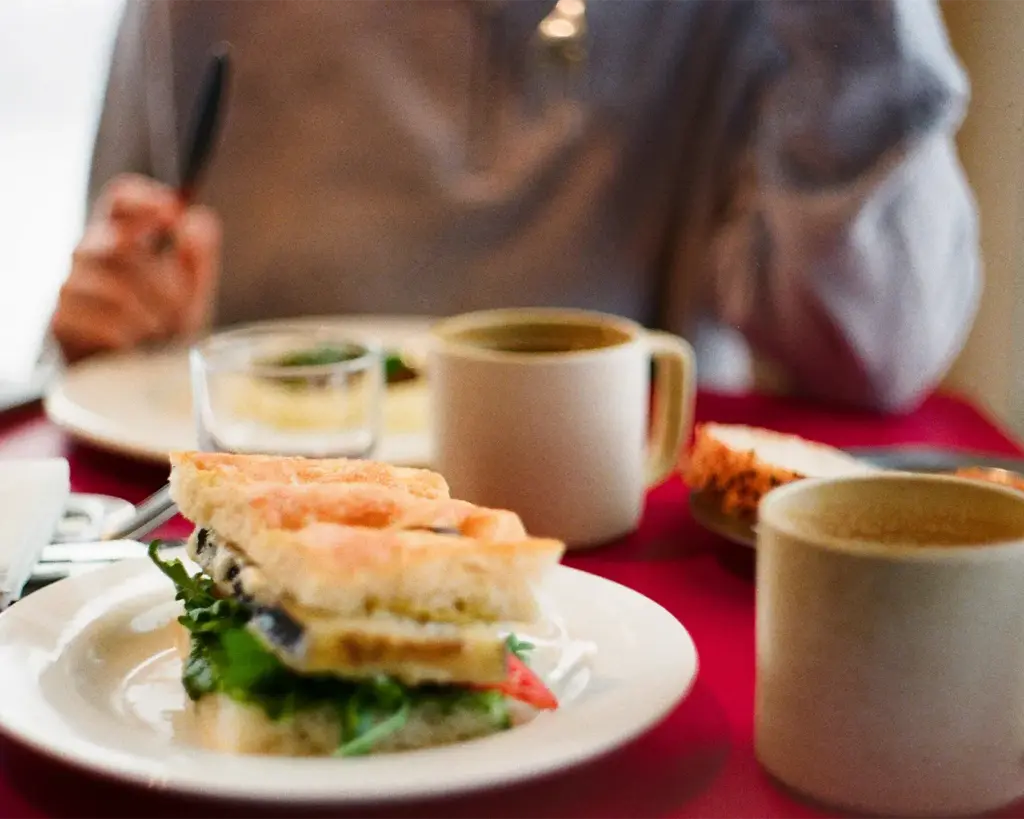 A close-up of a sandwich with greens and tomatoes on a white plate. Two coffee mugs and a blurred figure holding a fork are in the background, creating a cozy café vibe.
