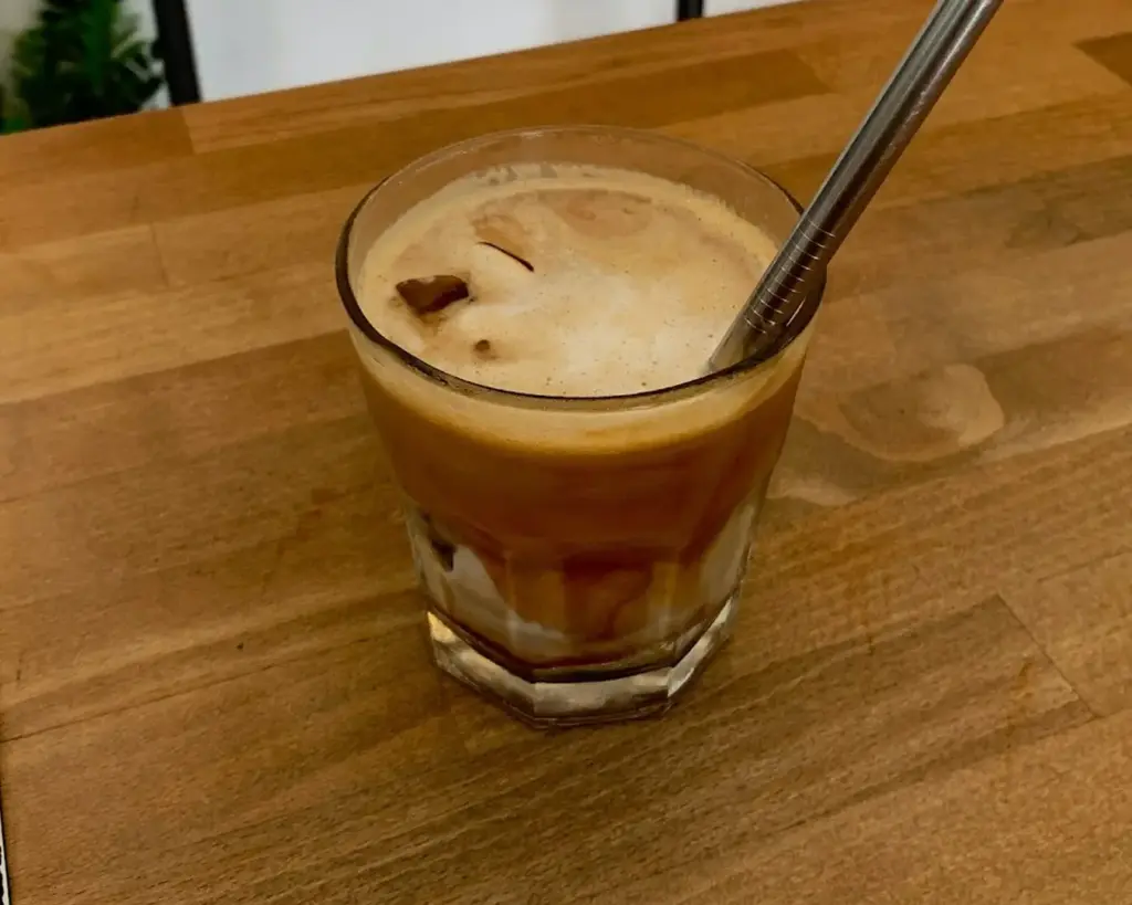 A glass of iced coffee sits on a wooden table, featuring creamy swirls and a metal straw. The setting conveys a cozy, inviting atmosphere.