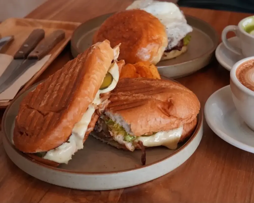 Grilled panini with melted cheese and pickles on a plate, alongside a burger, cappuccino, and cutlery set. Warm, inviting dining scene.