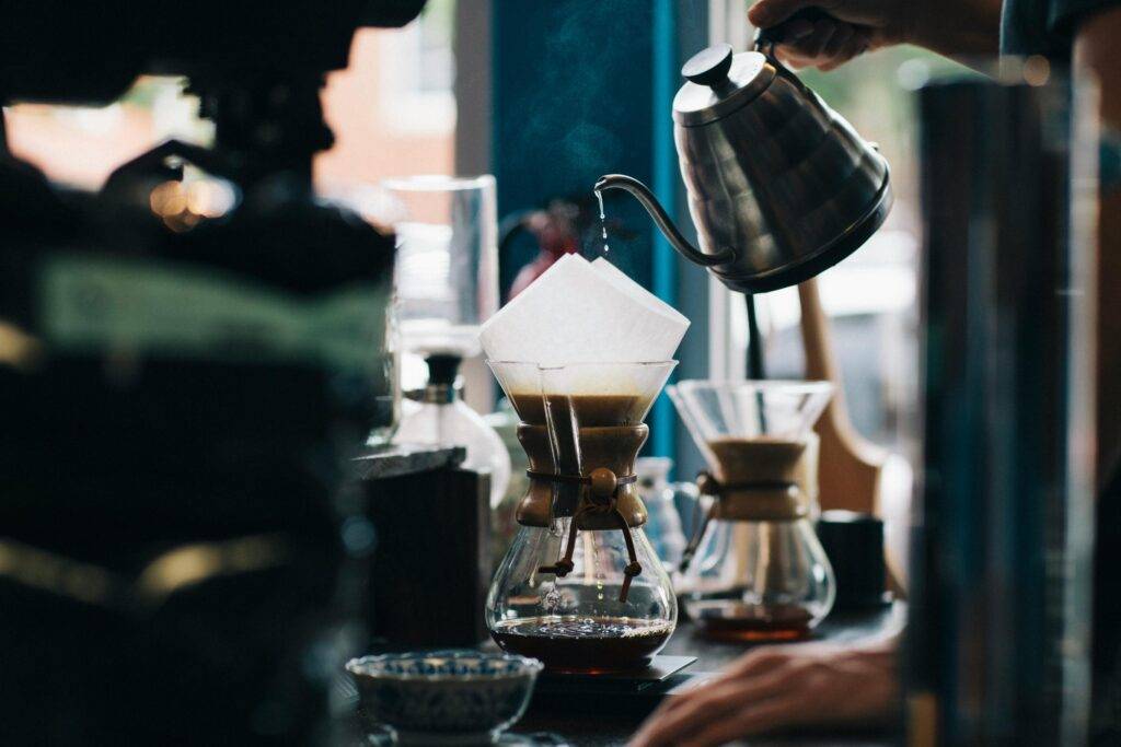 A barista carefully pours steaming water from a stainless steel gooseneck kettle into a glass pour-over coffee maker. The shot features a shallow depth of field and rich, moody tones that highlight the delicate stream of water and rising steam.