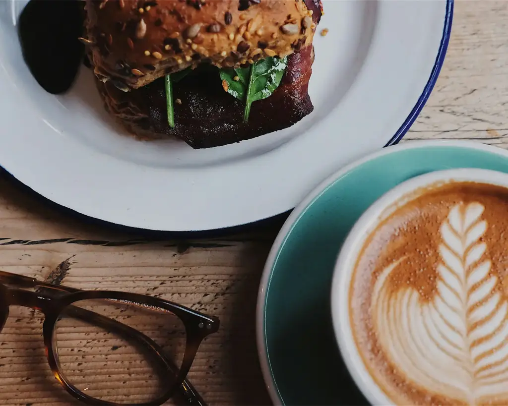 A close-up of a hearty sandwich with greens on a seeded bun beside a latte with intricate leaf latte art. Glasses rest nearby on a rustic table.