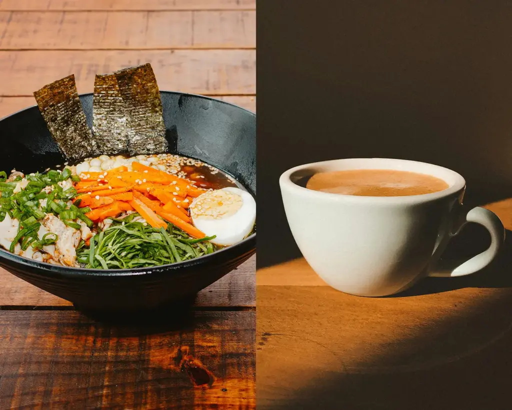 A black bowl of ramen with vegetables, egg, and seaweed sits beside a white cup of coffee on a wooden table, creating a cozy, inviting dining scene.
