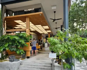 Modern café named "Alchemist" with wooden features, surrounded by lush greenery. Customers are ordering inside, creating a vibrant, inviting atmosphere.