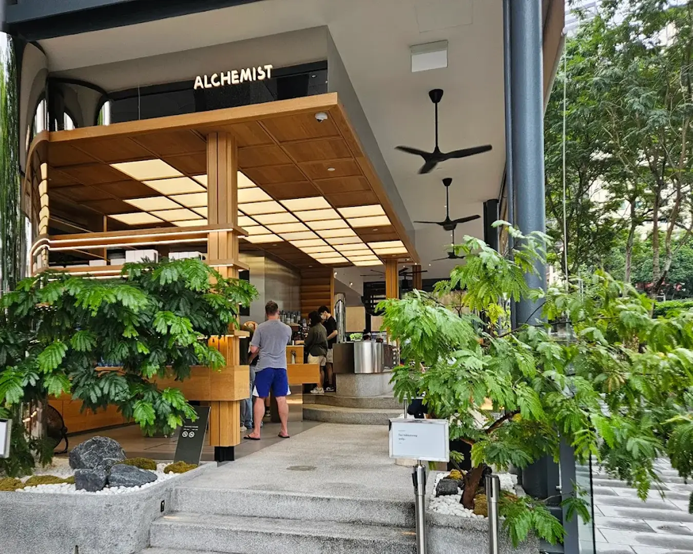 Modern café named "Alchemist" with wooden features, surrounded by lush greenery. Customers are ordering inside, creating a vibrant, inviting atmosphere.