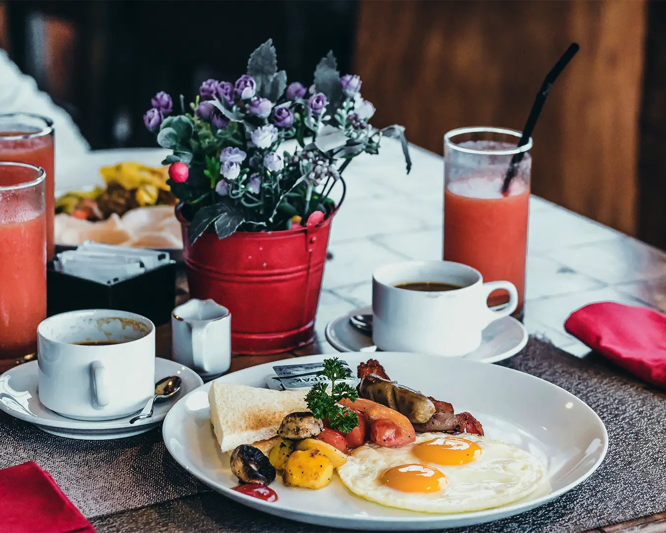 A cozy breakfast setting with a plate of sunny-side-up eggs, sausages, toast, and grilled vegetables. Nearby are coffee cups, a red fruit juice, and a vase of purple flowers.