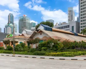 A traditional building with a sloped roof and lush greenery stands in front of modern skyscrapers under a blue sky, blending heritage with urban development.