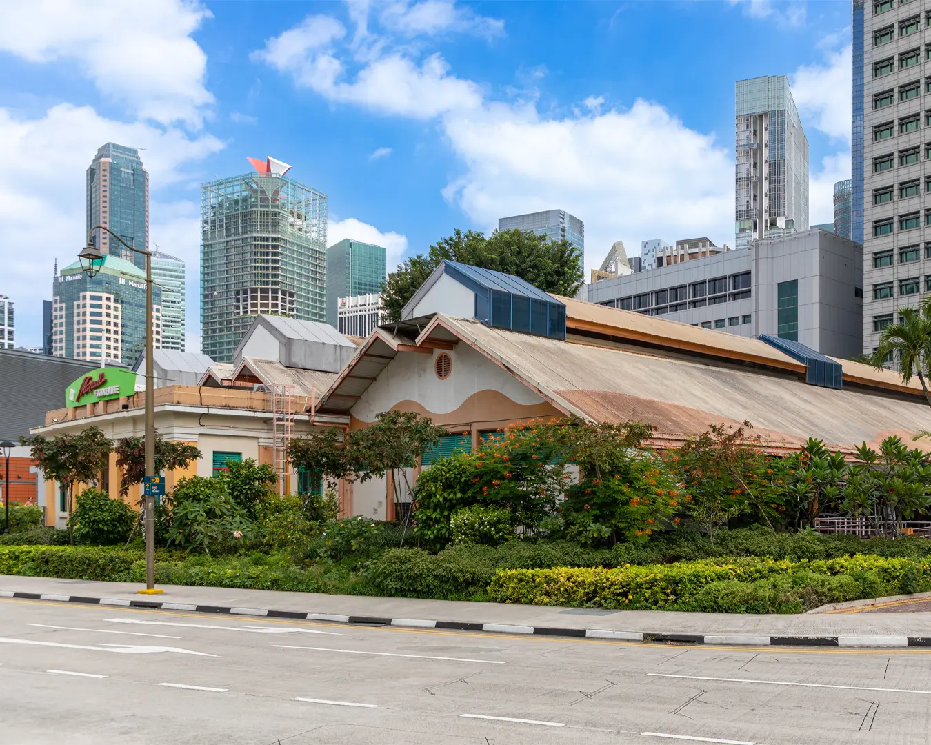 A traditional building with a sloped roof and lush greenery stands in front of modern skyscrapers under a blue sky, blending heritage with urban development.