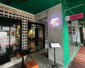 Exterior of a cafe with a modern, industrial look, featuring a glass door entrance, a neon sign reading "Brew Club," and brick and green elements.