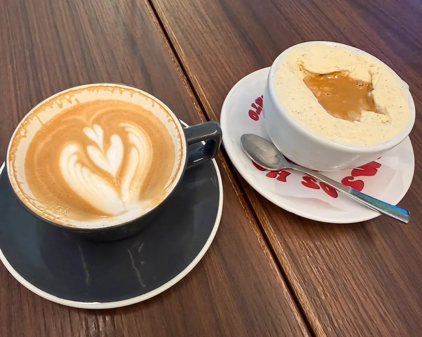 A latte with heart-shaped foam art in a dark cup sits beside a bowl of vanilla ice cream topped with caramel. Both rest on a wooden table, creating a cozy and inviting scene.