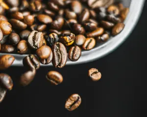 Close-up of coffee beans spilling from a white container against a dark background. The rich brown color and textures suggest freshness and aroma.