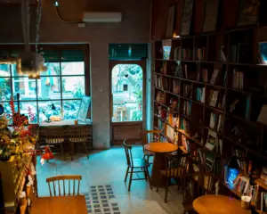 Cozy cafe interior with wooden tables and chairs, lined with bookshelves. Soft lighting creates a warm, inviting atmosphere. Large windows show lush plants outside.
